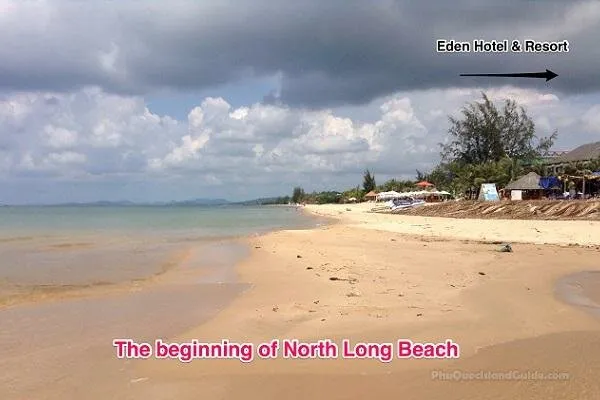 A wide shot of a sandy beach with calm ocean water under a cloudy sky. Buildings and tropical trees line the right side of the beach.