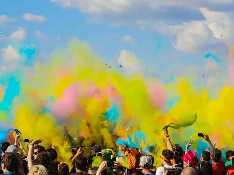 A crowd of people at an outdoor color festival, with vibrant yellow, pink, and blue powder filling the air against a blue sky. Many are holding up phones.