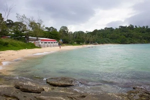 A tropical beach with clear turquoise water, a sandy shore, and lush green trees. A building with a red roof is visible on the left, and a few people are swimming.
