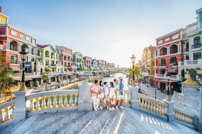 A Venice-style canal lined with brightly coloured shophouses, with a family standing on an ornate stone bridge