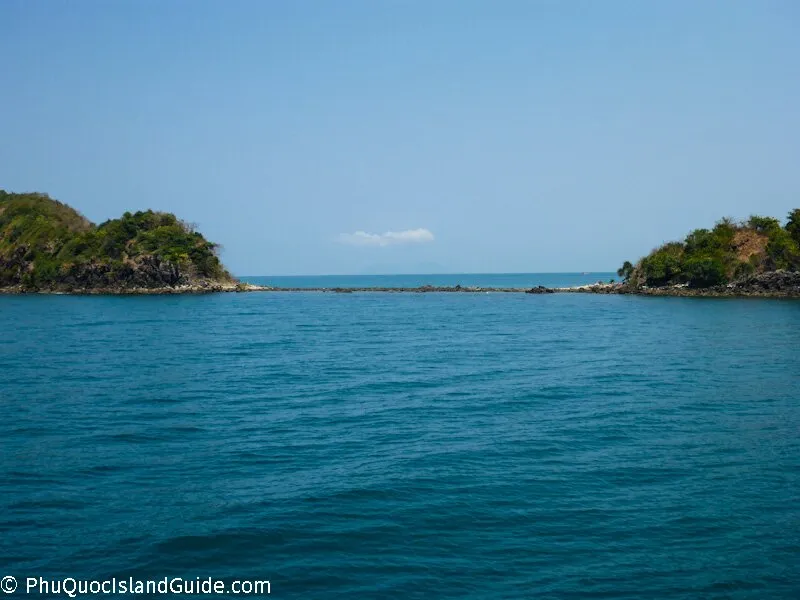 Two small, rocky islands covered in green vegetation, separated by a narrow channel of blue sea and connected by a low-lying rocky bar under a clear blue sky.