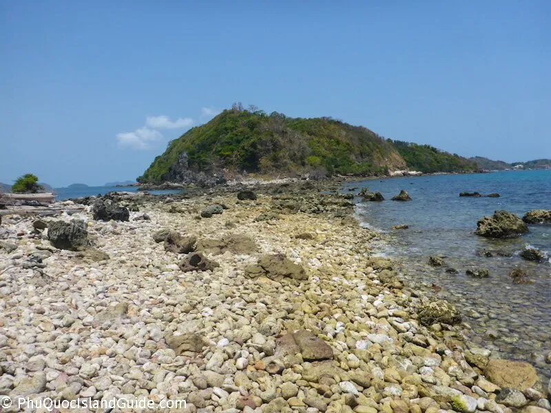 A rocky beach with clear blue water leading to a lush green island under a bright blue sky, with some small clouds.