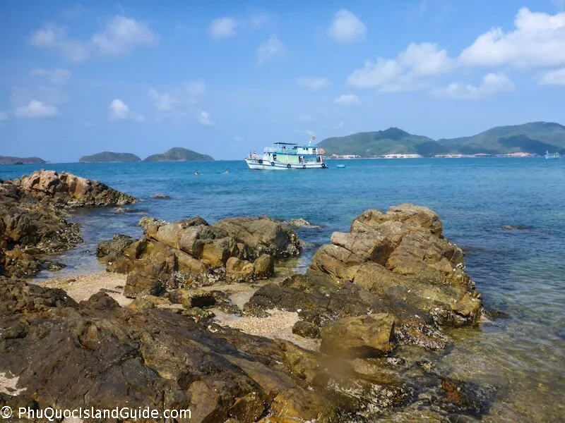 A scenic view of a rocky tropical coastline with clear blue water, a boat, and distant green islands under a bright blue sky with scattered clouds.
