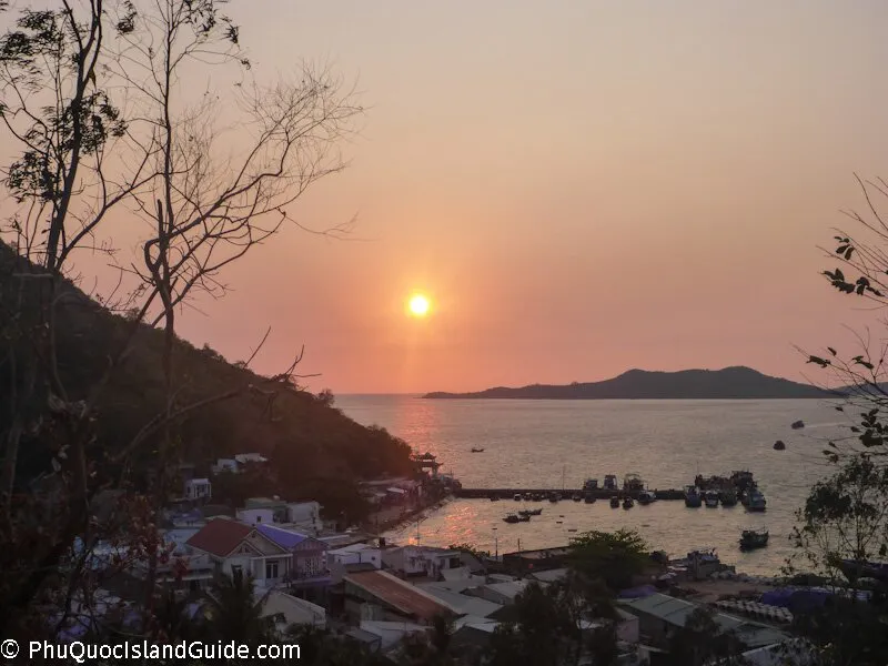 A scenic view of a sunset over a bay with a fishing village, a pier full of boats, and mountains in the background, seen from a hillside.