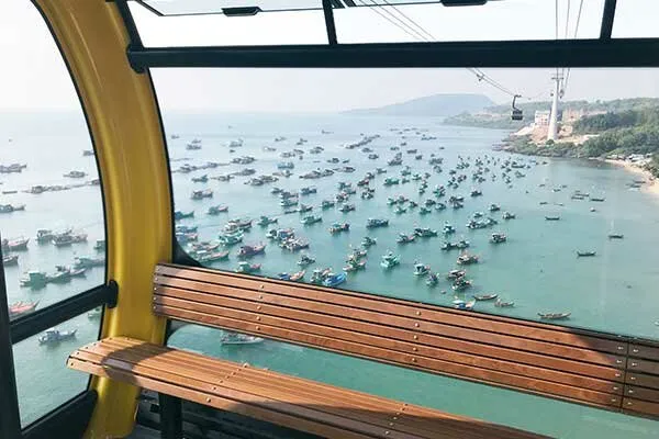 A view from inside a cable car cabin looking out over a bay filled with numerous fishing boats, with a coastline and hills in the distance.