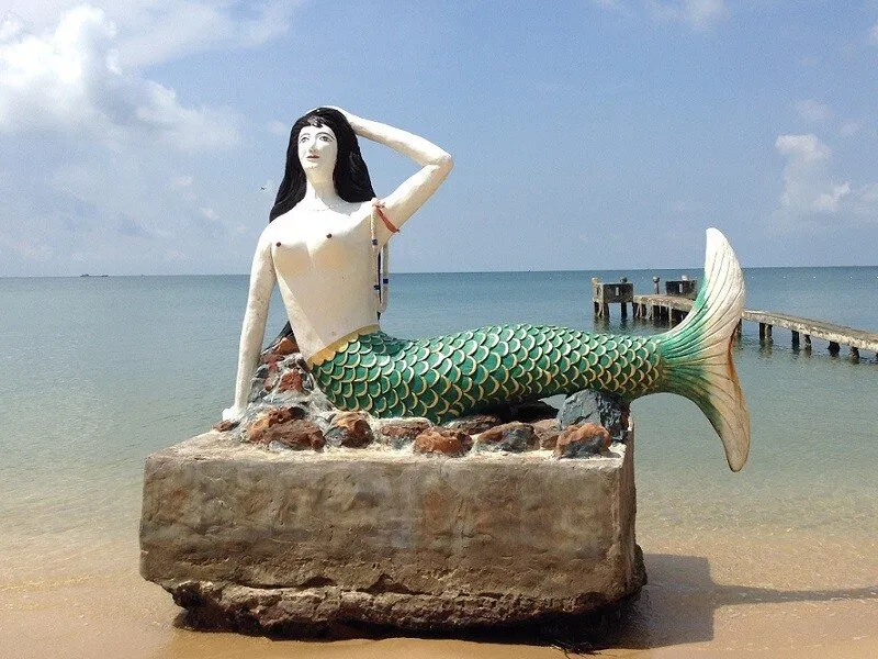 A mermaid statue with a green tail sits on a concrete base on a sandy beach. The ocean and a wooden pier are visible in the background under a blue sky.