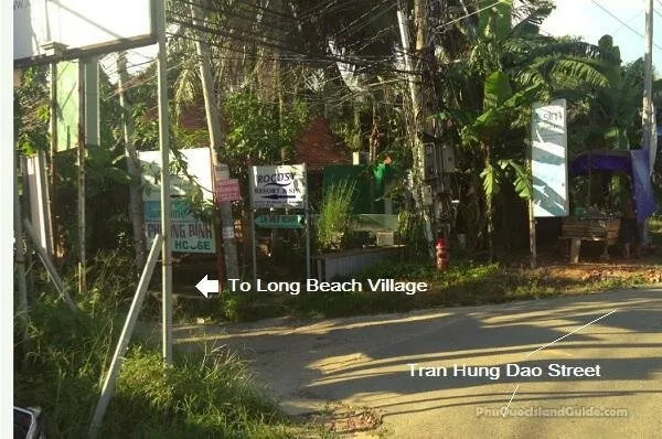 Roadside junction on Tran Hung Dao Street with guesthouse signboards, an arrow labelled “To Long Beach Village” pointing down a side lane