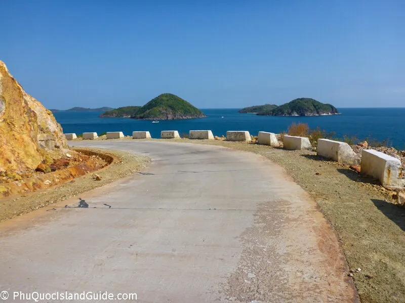 A winding coastal road with concrete barriers, overlooking green islands in a blue sea under a clear sky.
