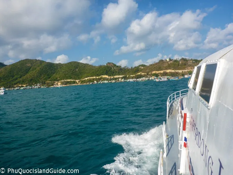 View from a boat moving through blue water, with a green, hilly coastline and a village in the background under a cloudy sky.
