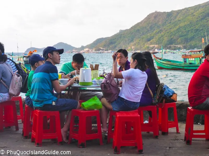 A group of people eating at an outdoor restaurant by the sea, sitting on red plastic stools. Fishing boats are visible in the water, with green mountains in the background.