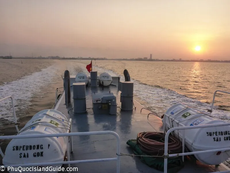 View from the stern of a boat at sunset, showing the wake, distant city skyline, and life rafts on deck. A Vietnamese flag flies at the back.