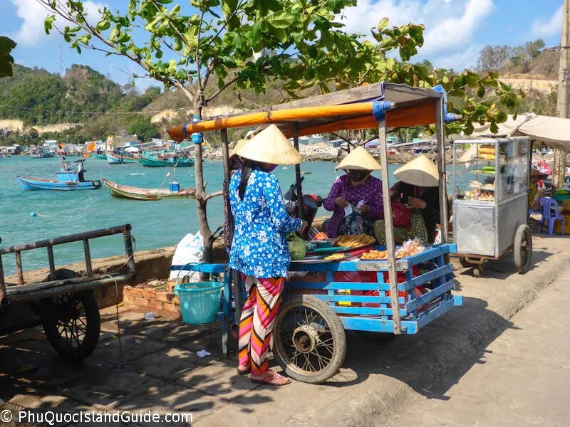 Three women in conical hats sell food from carts by the sea, with fishing boats and a green hillside in the background under a clear sky.