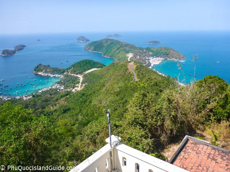 Panoramic view of a tropical bay with numerous small islands, lush green hills, a coastal village, and many boats anchored in the turquoise water.