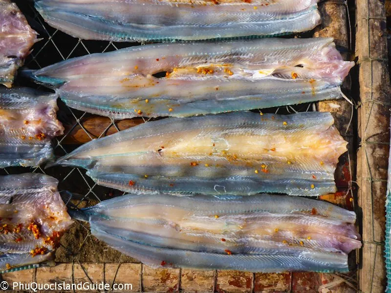 Close-up of several flattened, dried fish fillets, possibly seasoned with chili flakes, laid out on a mesh rack for sun-drying.