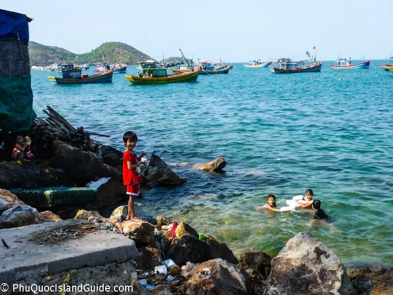 Children playing in the sea and on a rocky shore with fishing boats anchored in the background under a clear sky. Some debris is visible on the shore.