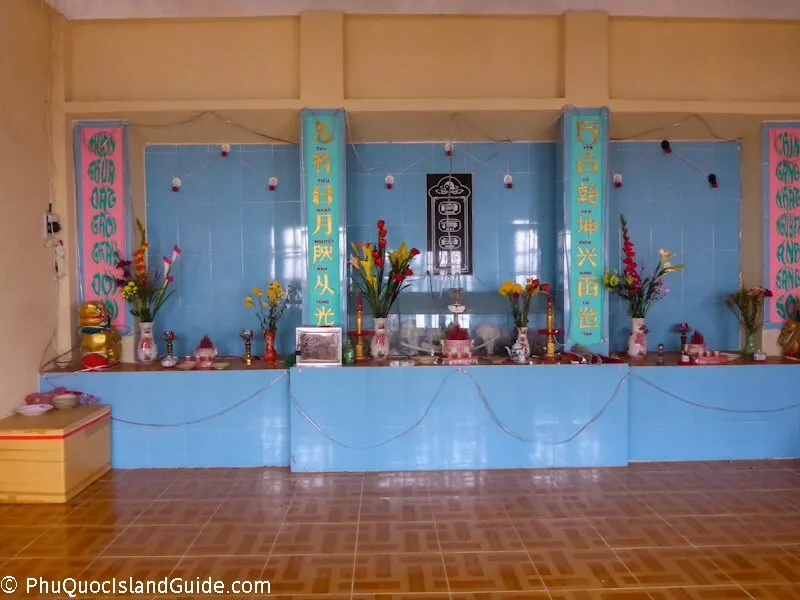 An indoor altar or shrine with blue tiled walls, decorated with vases of colorful flowers, offerings, and banners with Vietnamese text. A wooden box is on the left.
