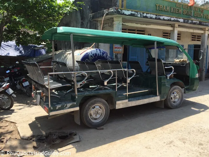A green open-sided shuttle bus with multiple rows of seats is parked on a dirt road next to a building with Vietnamese text.