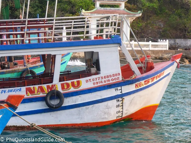 A close-up of the bow of a white, red, and blue boat named “NAM DU” docked in clear blue water, with another boat and a traditional white structure in the background.