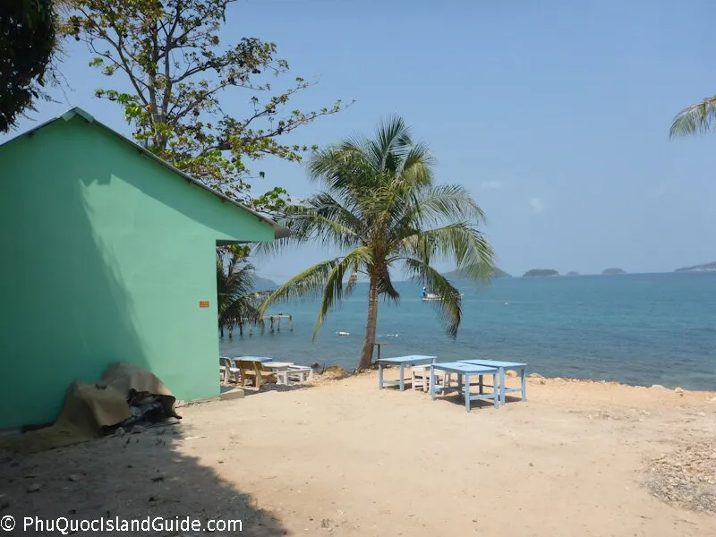 A green building next to a sandy area with blue tables, a palm tree, and the clear blue sea with distant islands under a bright sky.