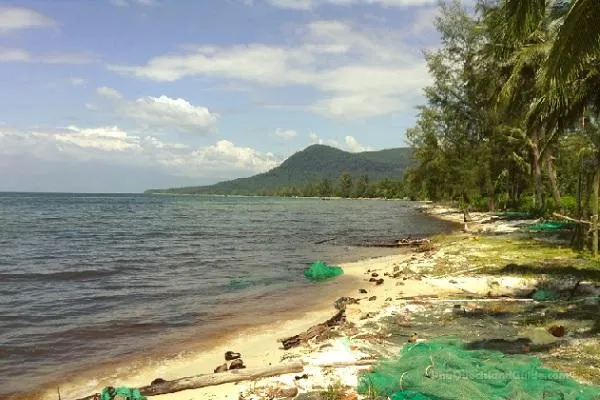 A tropical beach with calm ocean water, green fishing nets on the sand, lush green trees, and mountains under a blue sky with white clouds.