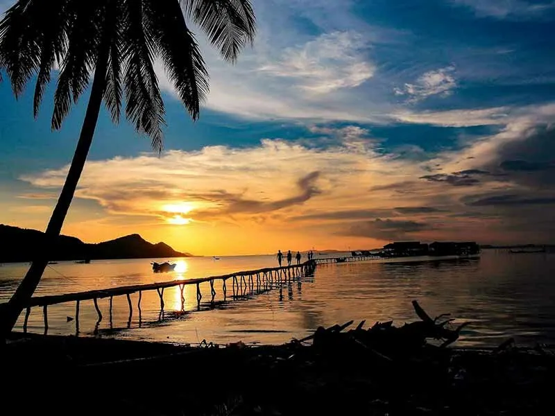 A vibrant sunset over a tropical sea, with a long wooden pier extending into the water. Silhouetted people walk on the pier, and a palm tree frames the left side.