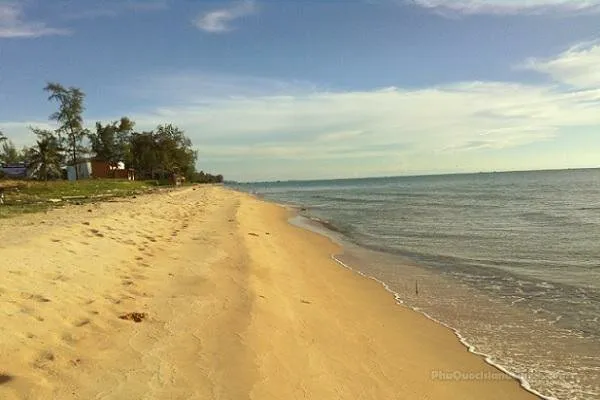A wide shot of a sandy beach with gentle waves lapping the shore. Green trees and small structures line the distant coastline under a blue sky with scattered clouds.
