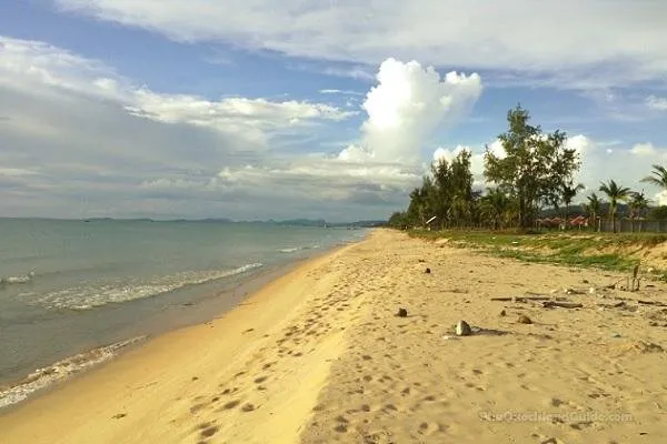 A sandy beach with gentle waves, the ocean, and a line of green trees along the shore under a cloudy sky. Footprints are visible in the sand.