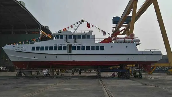 A white and red ship, possibly a ferry, on dry land in a shipyard, with large yellow cranes in the background. Flags are strung across the ship’s top deck.