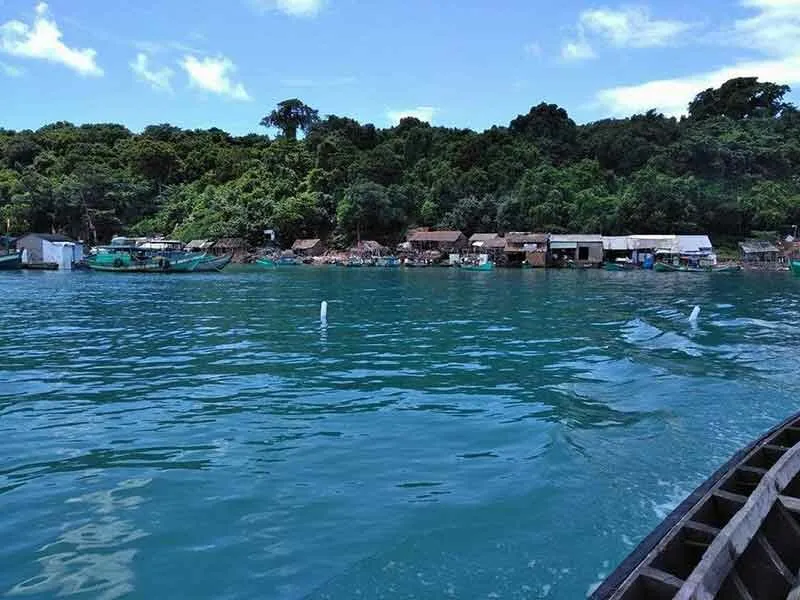 A view from a boat showing a tropical coastline with dense green trees, small buildings, and fishing boats docked along the shore under a blue sky.