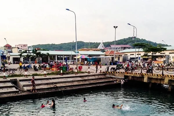 A bustling waterfront scene with people swimming in the foreground, buildings, and a green, forested hill in the background.
