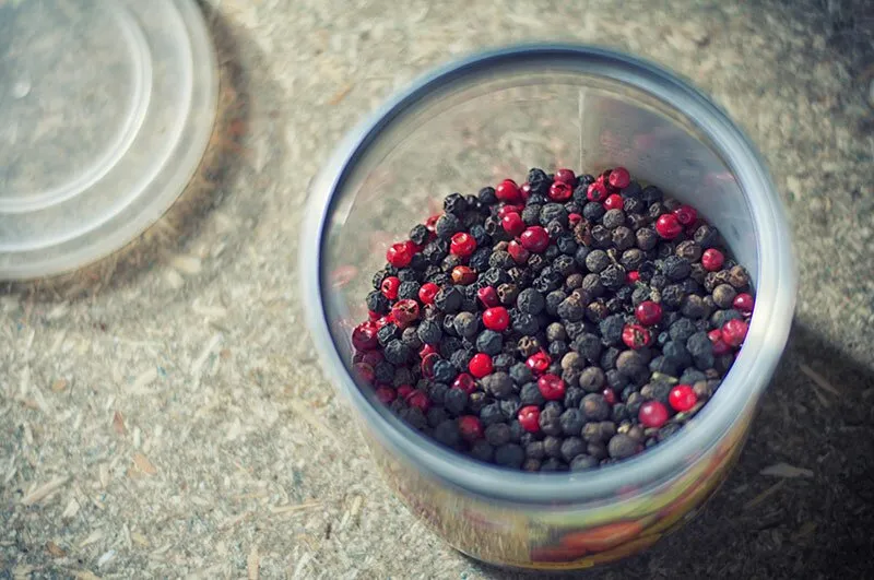 A clear plastic container filled with a mix of black and red peppercorns, with a plastic lid next to it on a textured surface.