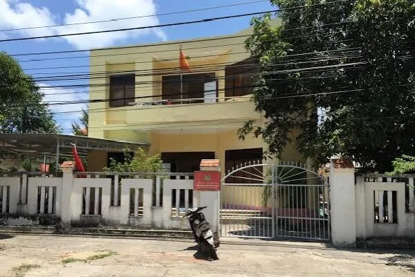 A yellow two-story building with a fence, a closed gate, and a scooter parked on the street. A Vietnamese flag hangs from the balcony. Trees and power lines are visible.