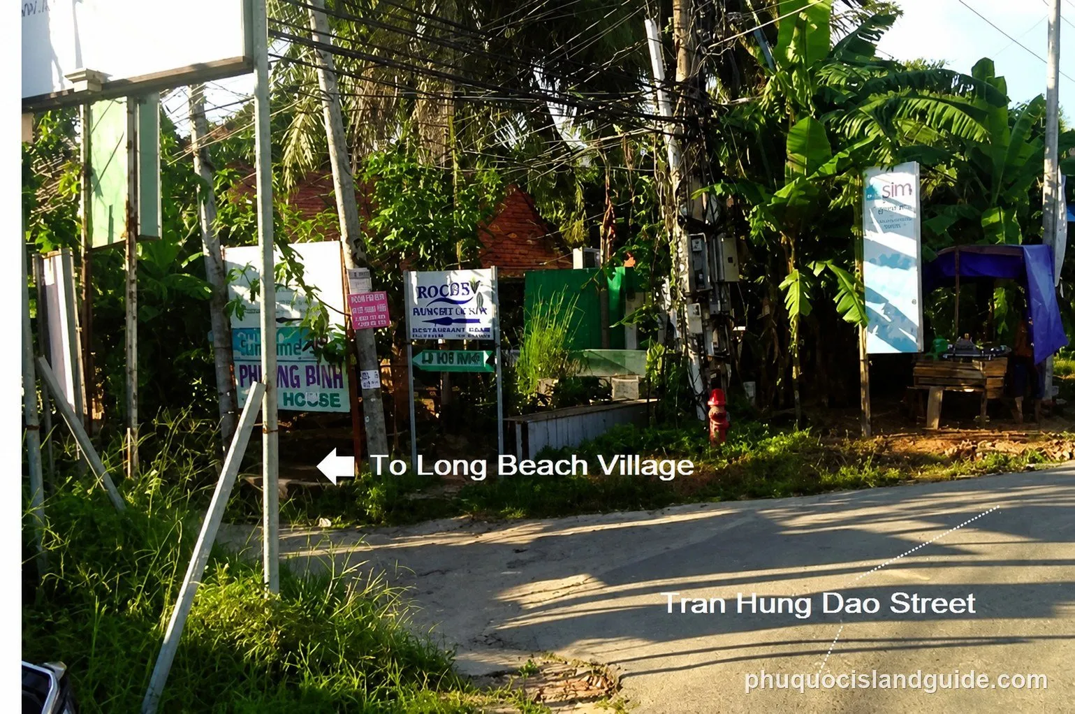 Roadside junction on Tran Hung Dao Street with guesthouse signboards, an arrow labelled “To Long Beach Village” pointing down a side lane