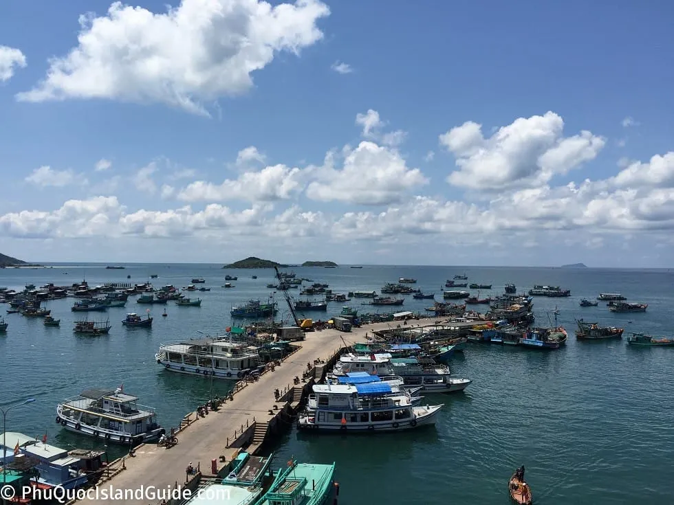 An aerial view of a busy harbor filled with numerous fishing boats and passenger vessels docked at a long pier under a bright blue sky with white clouds. Distant islands are visible.