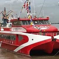 A red and white catamaran high-speed ferry boat docked in a port, with flags flying from its mast. Cranes and industrial structures are visible in the background.