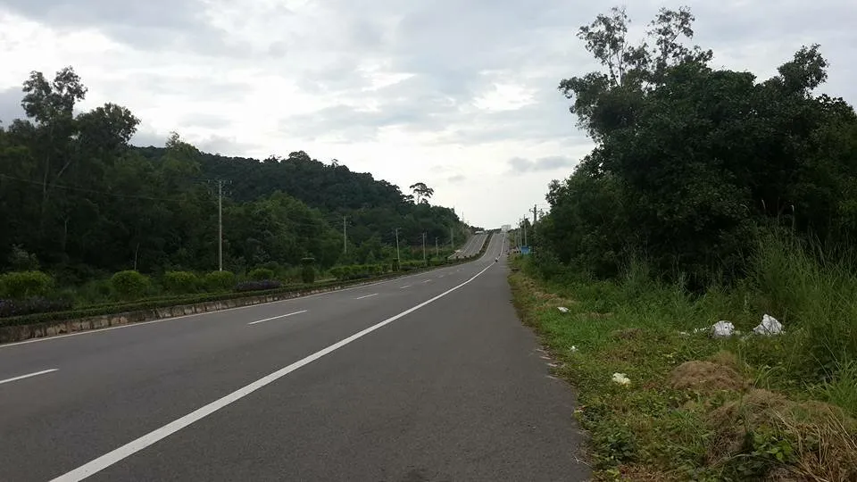 A long, winding highway stretches through a lush green, hilly landscape under a cloudy sky. Dense tropical trees line both sides of the road.
